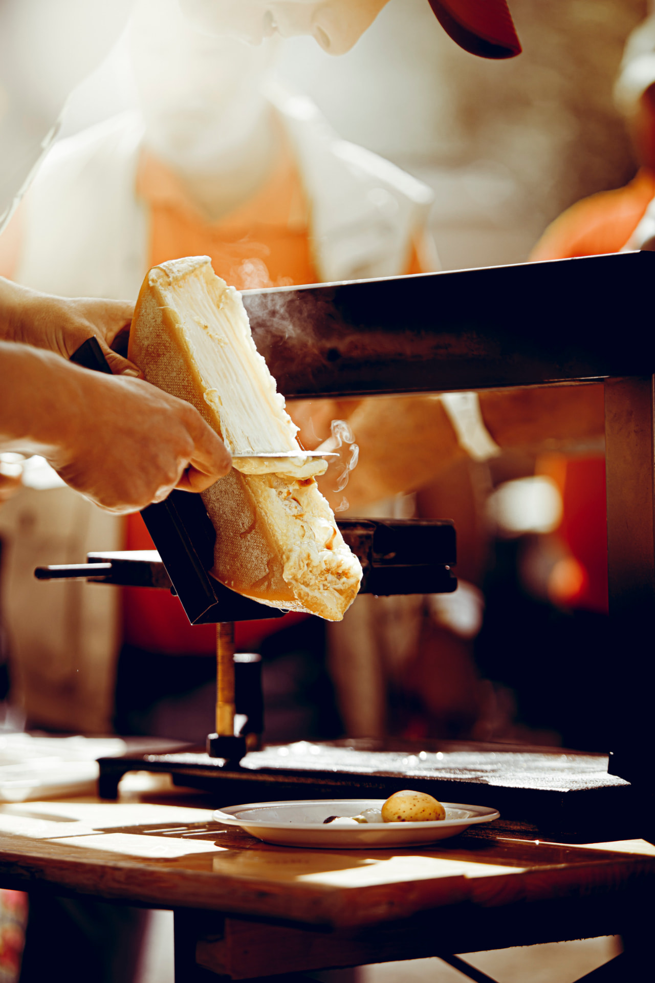 Demi-meule de fromage fondue devant un four à raclette, avec une pomme de terre sur une assiette